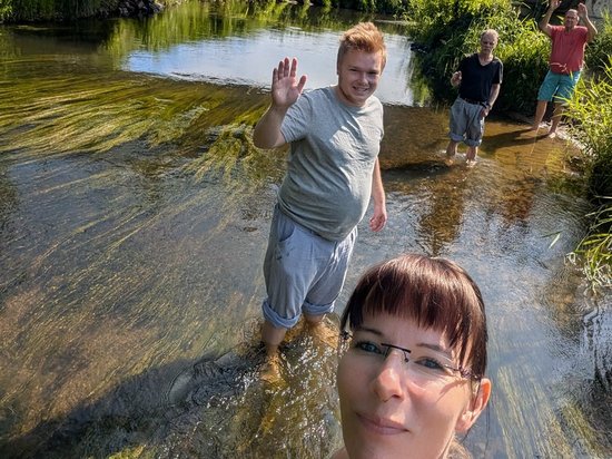 Eine Frau macht ein Selfie im flachen Fluss. Hinter ihr winkt ein Mann fröhlich in die Kamera. Zwei weitere Personen stehen weiter hinten im Wasser. Auf einer Brücke darüber beobachten vier Personen die Szene von oben.