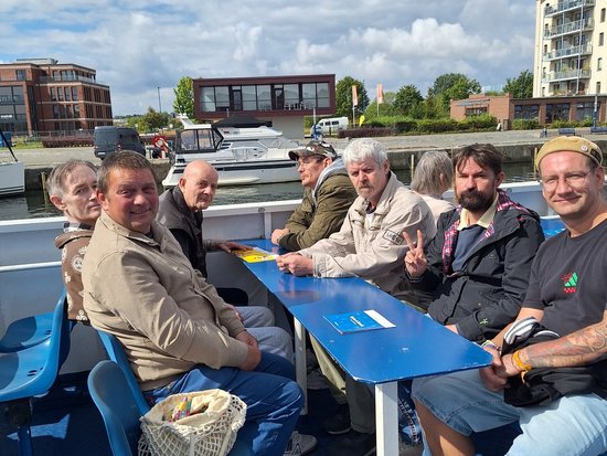 Eine Gruppe von acht Personen sitzt entspannt an einem blauen Tisch auf dem Deck eines Bootes. Im Hintergrund sind städtische Gebäude und ein blauer Himmel mit Wolken zu sehen. Die Atmosphäre wirkt gesellig und freundlich.