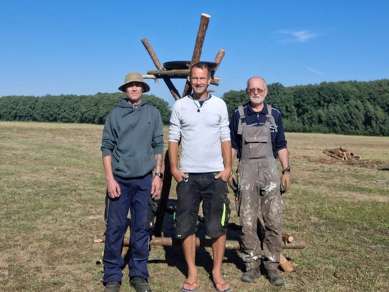Drei Männer stehen auf einer Wiese vor einem klaren blauen Himmel. Hinter ihnen befindet sich eine hölzerne Konstruktion. Der Mann links trägt einen Hut, der mittlere ein weißes Oberteil und der rechte eine Arbeitslatzhose. Im Hintergrund ist eine Baumreihe zu sehen.
